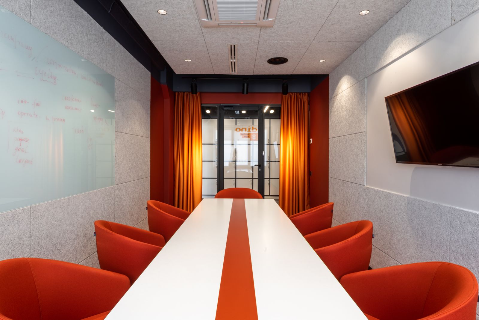 Set of comfortable orange armchairs placed around long white table in modern workspace with TV on wall and glass door