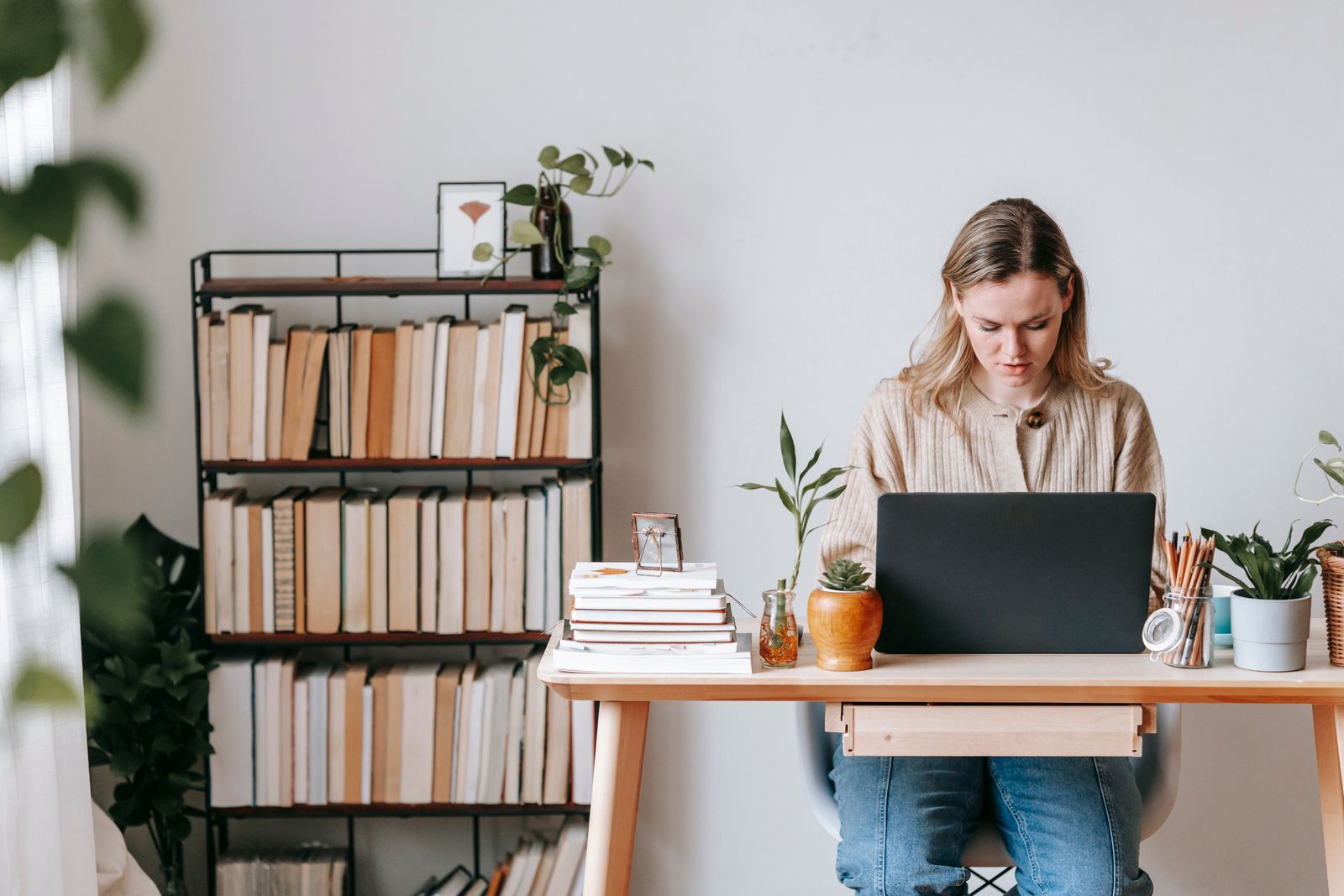 Focused female remote employee working on project on netbook at table with potted plants against bookcase at home on light background